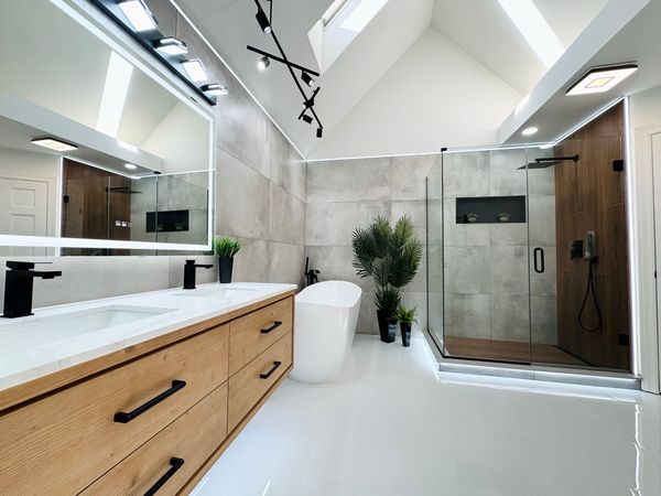 Primary bathroom after, with a wood-tone vanity with black pulls, a large lit mirror and a glass shower